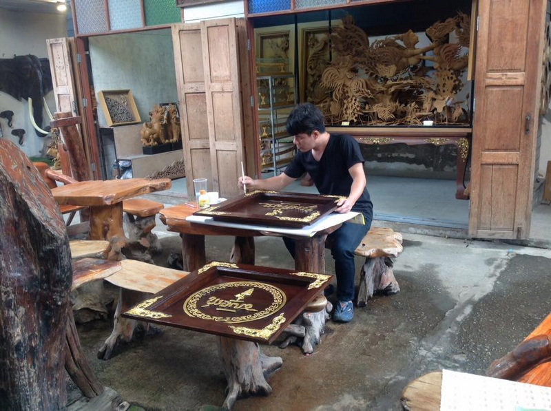 A person carving wood at a workshop with wooden carvings and furniture around at Baan Tawai.