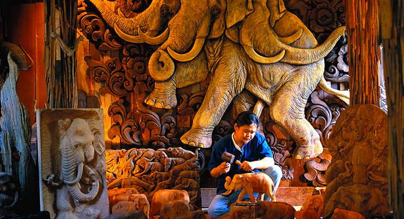 Wood Carving Workshop Scene at Baan Tawai Wood Carving Village Person sitting among detailed wooden carvings and sculptures inside a workshop at Baan Tawai Wood Carving Village.