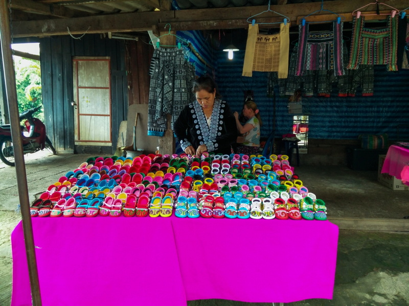 Woman arranging colorful slippers on a pink table inside a wooden shop with hanging fabrics at Hmong Doi Pui Village.