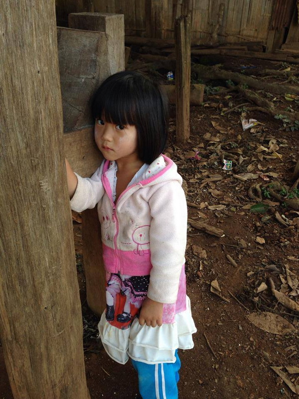 Young Girl in Nature Setting at Khun Chang Kian Village Young girl standing outdoors next to a wooden post with dirt ground and wooden structures behind her at Khun Chang Kian Village.
