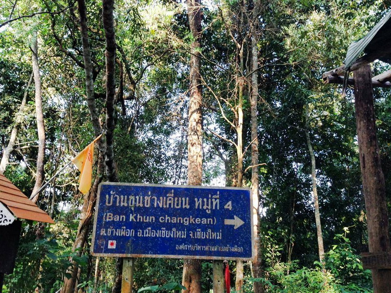 Sign to Ban Khun Chang Kian Blue sign indicating Ban Khun Chang Kian surrounded by tall trees and greenery
