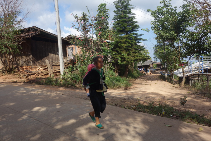 Woman carrying a child walking along a dirt road with trees and houses nearby at Khun Chang Kian Village.