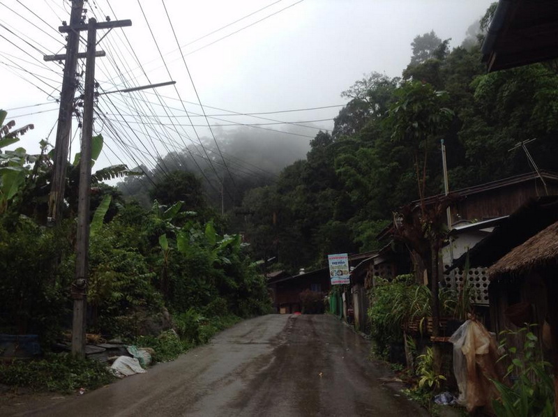 Rural Village Road in Fog at Mae Kanpong Village A rural road surrounded by small houses and dense greenery with a foggy hillside in background at Mae Kanpong Village.