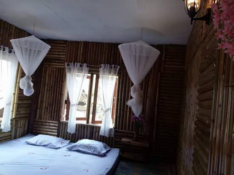 Interior of a wooden-themed bedroom with a bed and white curtains on windows at Ban Mae Kanpong.