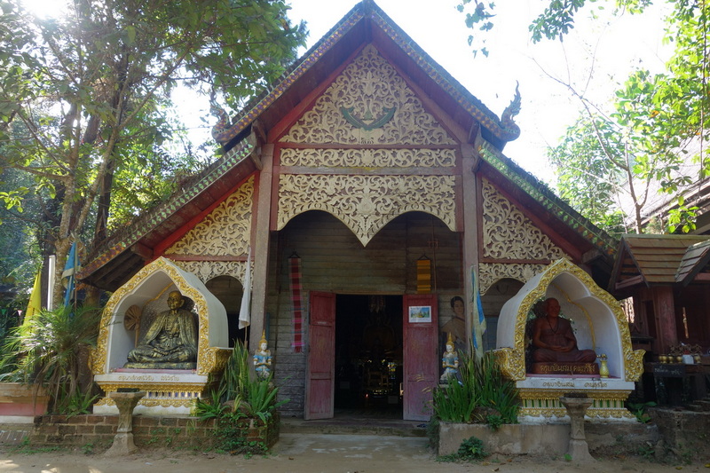 A wooden temple with carvings and statues surrounded by trees at Mae Kanpong Village.
