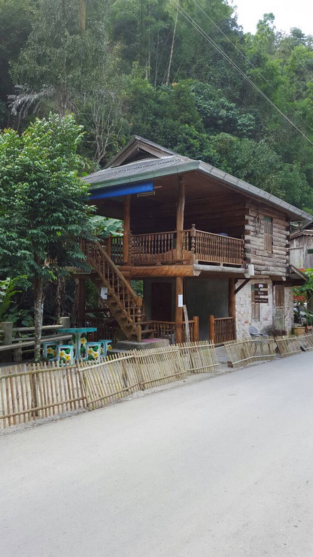 Two-story wooden house with balcony and staircase next to lush trees at Ban Mae Kanpong.