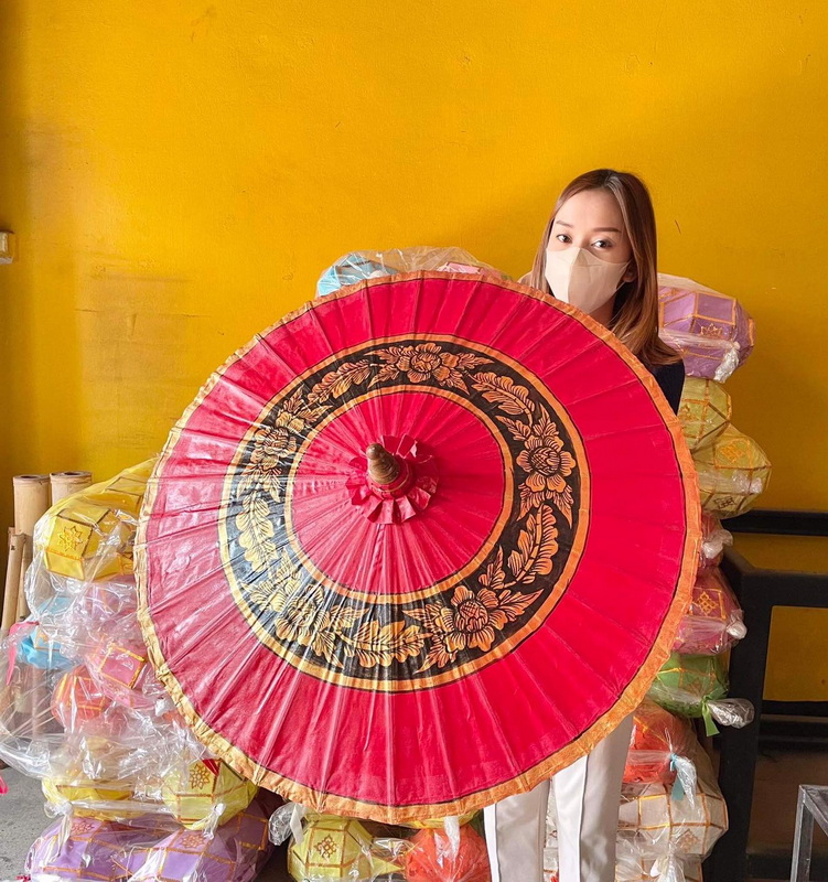 Woman with face mask holding a red and gold umbrella indoors against a yellow wall at San Kamphaeng Village.