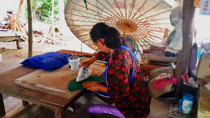 Woman Ironing Fabric at San Kamphaeng Village Woman ironing fabric under a traditional umbrella in a rustic workspace at San Kamphaeng Village.