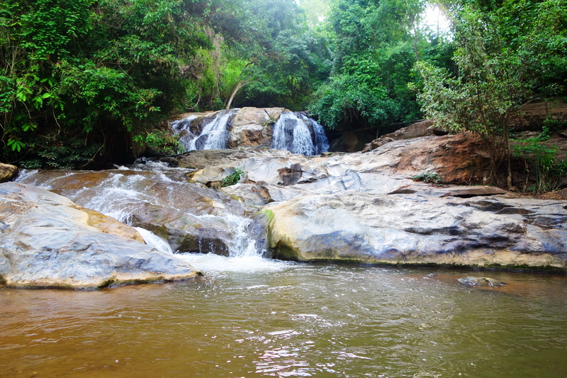 Water flowing over rocks into a pool surrounded by green trees and plants at Mae Sa Waterfall.
