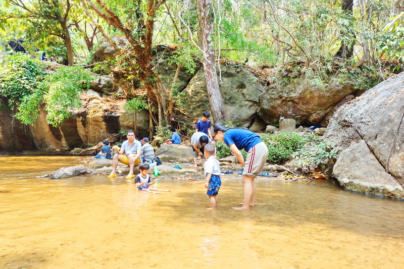 People enjoying a shallow river surrounded by trees and rocks in nature at Mae Sa Waterfall.