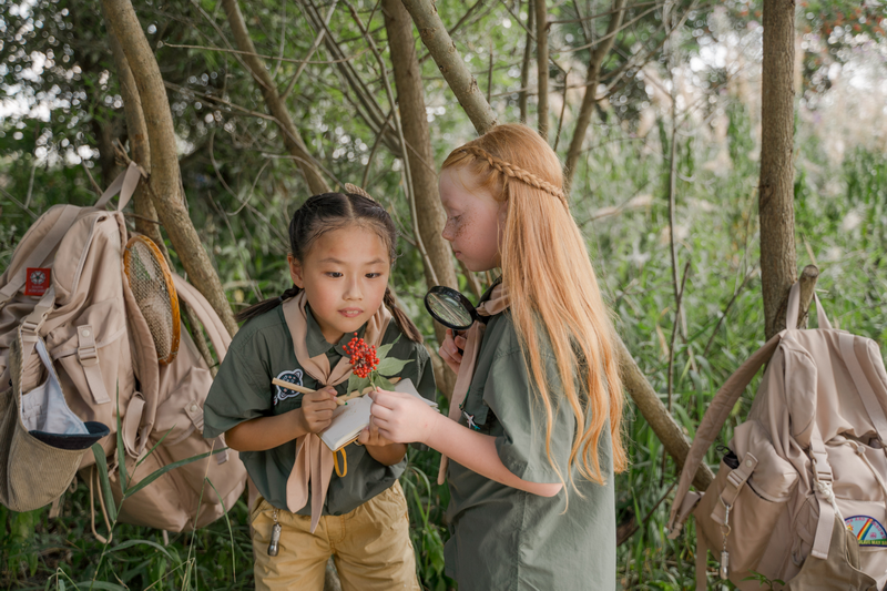 Two girls in scout uniforms studying a flower and leaf in a forested area at Nature Camp For Kids Chiang Mai.