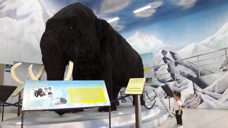 Child observing a large woolly mammoth replica in a museum with mountain murals at Chiang Mai Zoo.