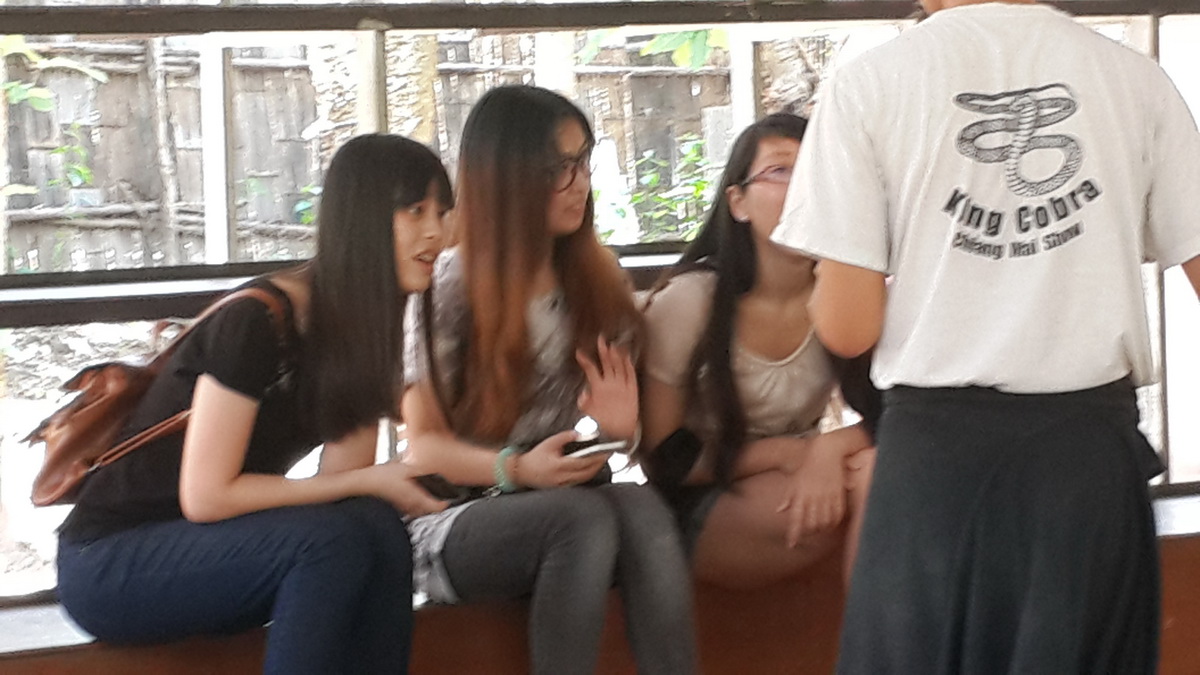 Three women sitting indoors on a bench talking to a person in a white shirt with snake logo at Mae Rim Snake Farm.
